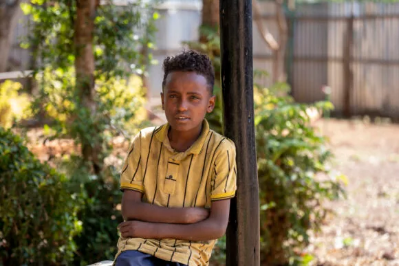 Poika nojaa ulkona katoksen tolppaan, päällään keltainen pikeepaita.