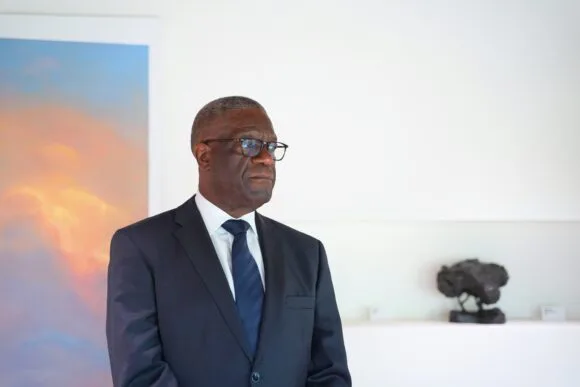 Pukuun pukeutunut mieshenkilö, Denis Mukwege, valkoisen seinän edessä.