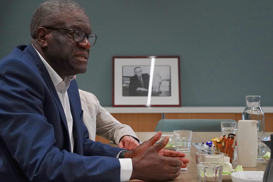 Tohtori Mukwege kokoushuoneen pöydän ääressä ja taka-alalla valokuvataulu toisesta rauhannobelistista, presidentti Ahtisaaresta.