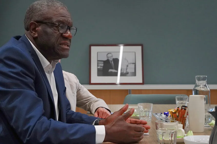 Tohtori Mukwege kokoushuoneen pöydän ääressä ja taka-alalla valokuvataulu toisesta rauhannobelistista, presidentti Ahtisaaresta.