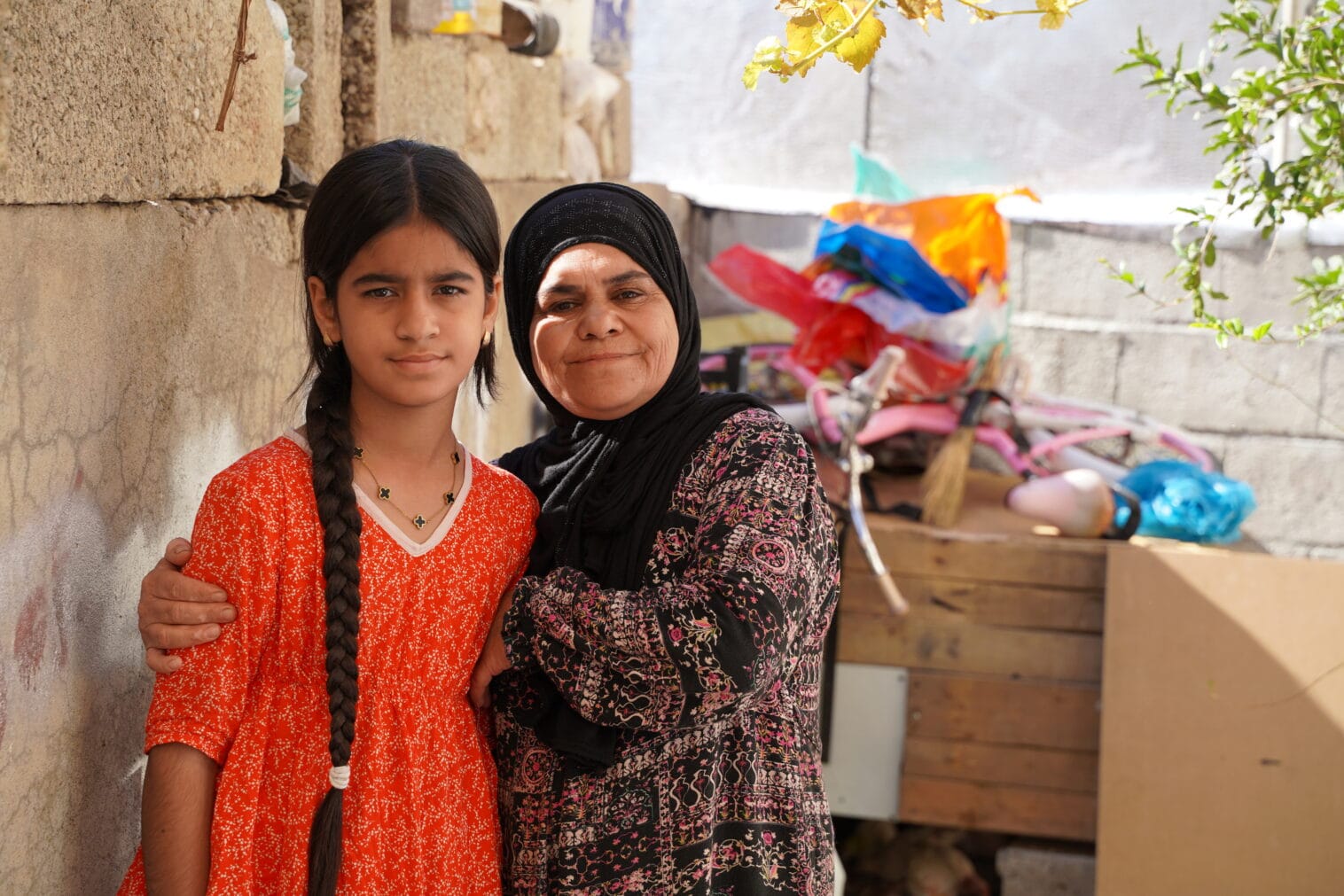 Kuulovammainen tyttö punaisessa mekossa. Äiti pitää kättään tytön ympärillä ulkona talon edessä.