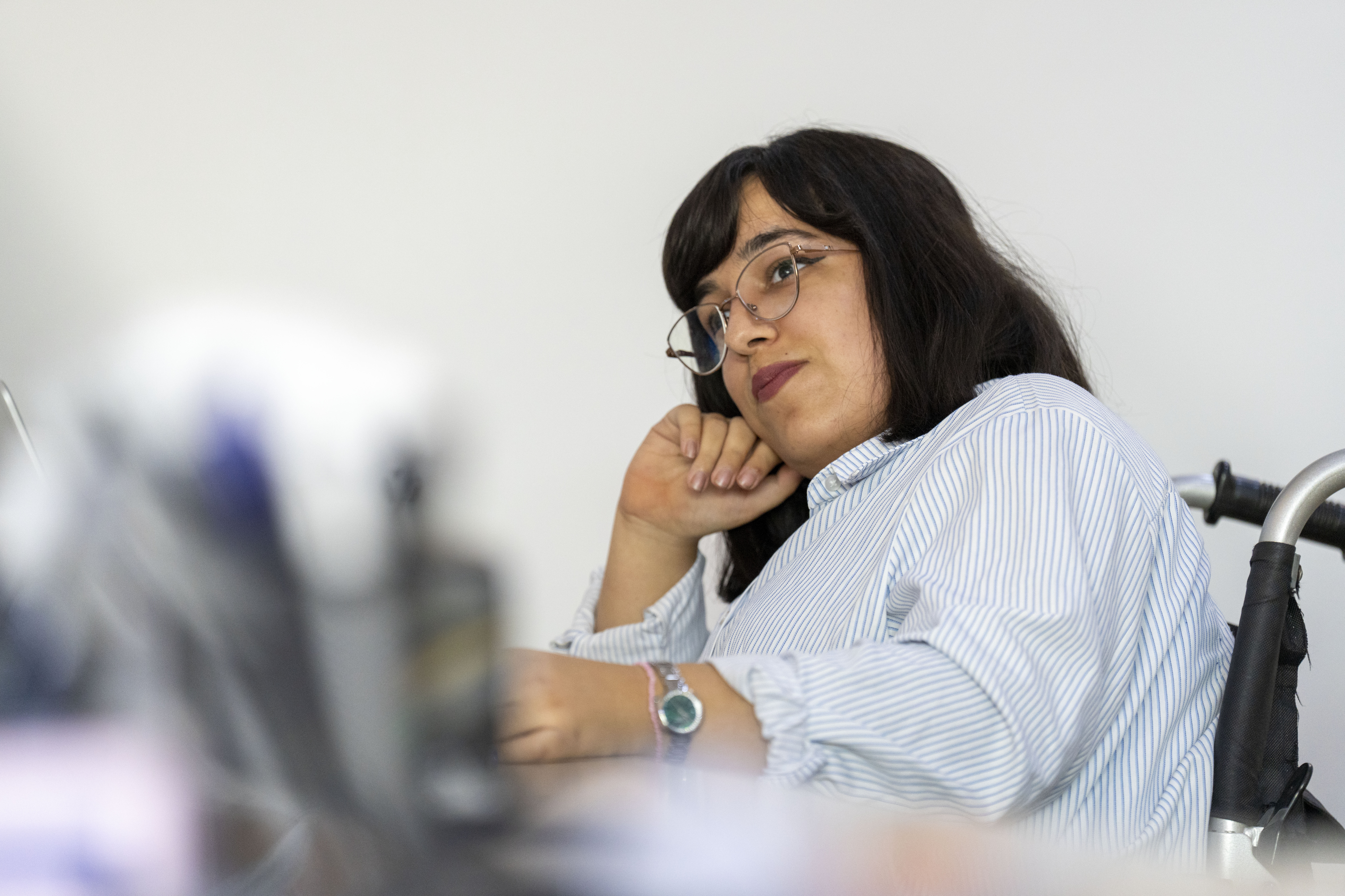 Pyörätuolissa istuva nuori nainen on kokouspöydän ääressa ja nojaa päätään toiseen käteensä.