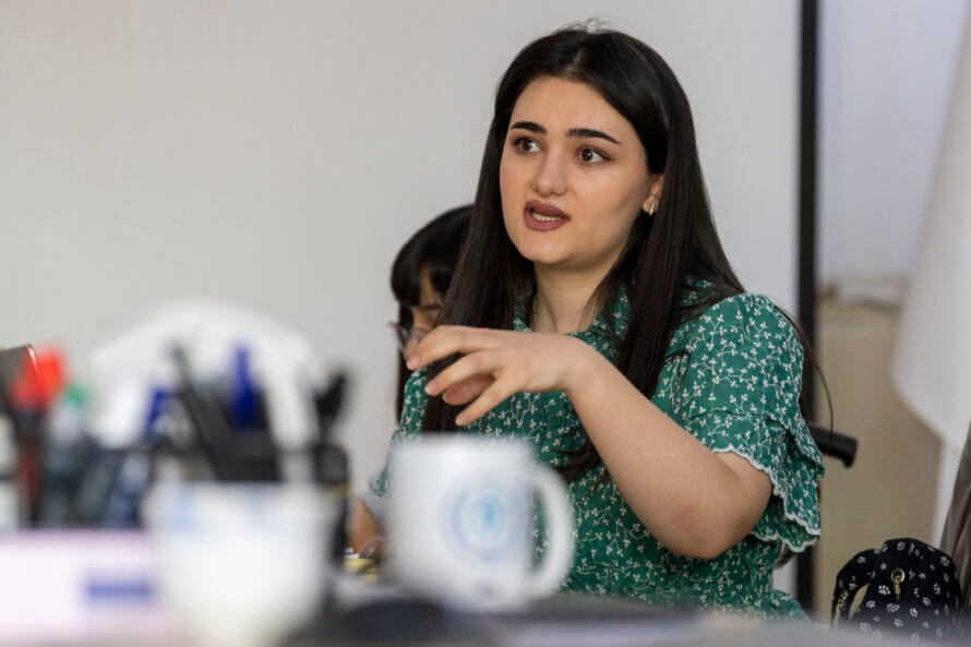 Vihreään mekkoon pukeutunut nuori nainen istuu pöydän ääressä ja puhuu.