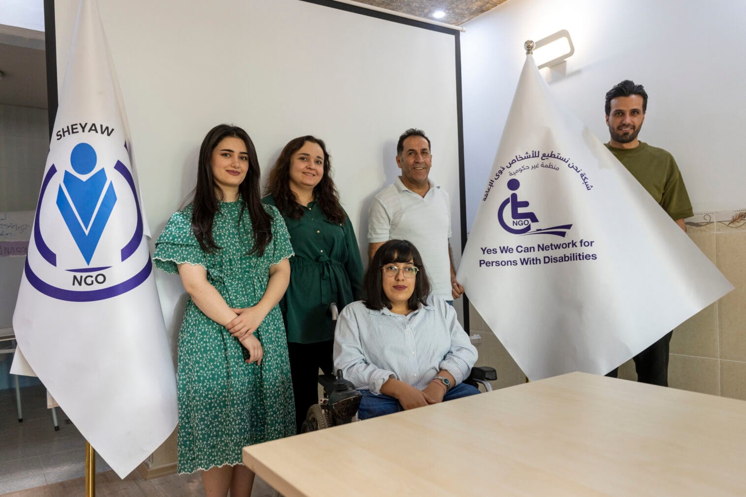 Neljä Sheyaw-järjestön työntekijää seisoo lippujen välissä, joissa on järjestön logo. Viides henkilö pitelee lippua niin, että logo ja teksti näkyvät. Hän on Fidan työntekijä.