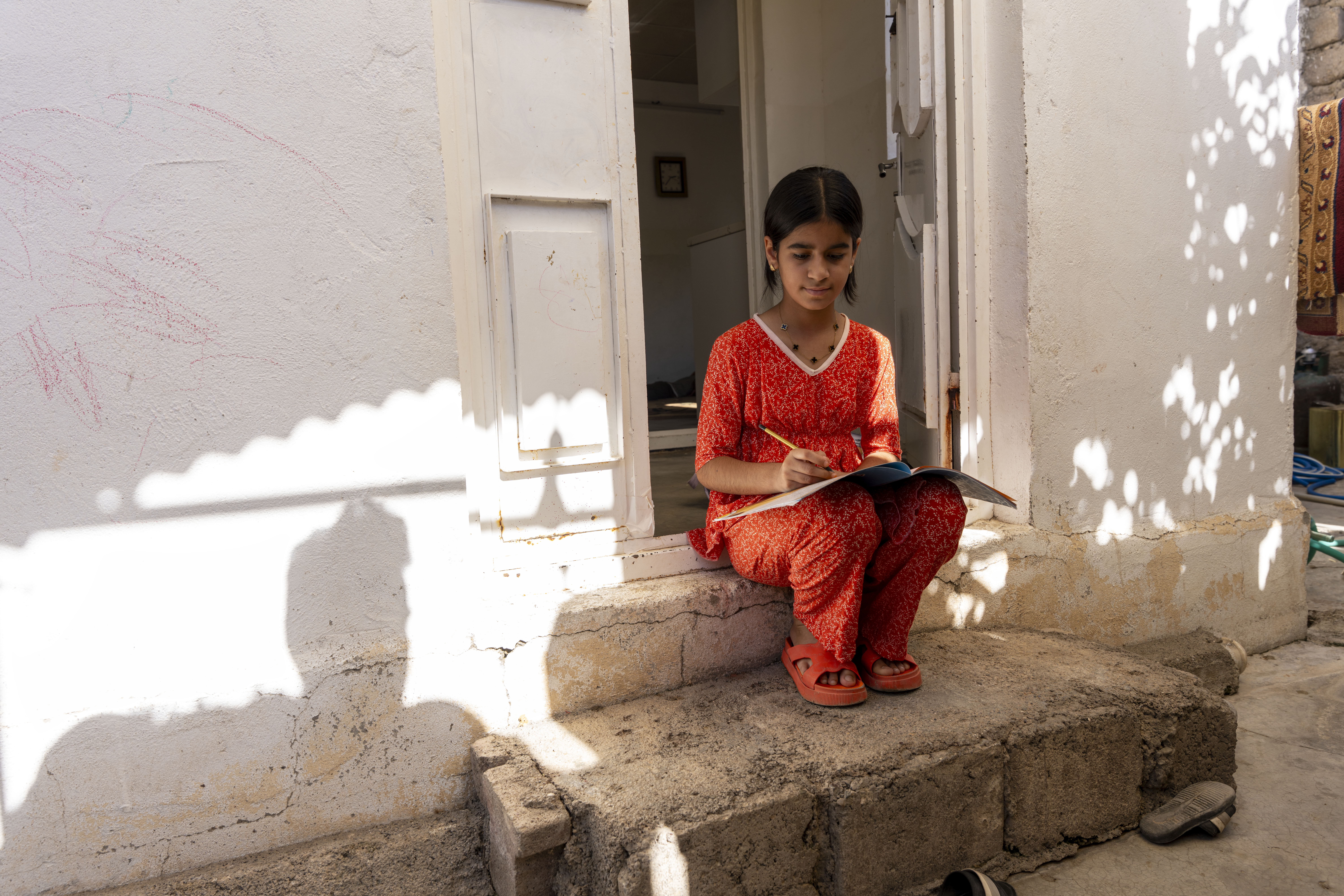 Tyttö istuu kotioven kynnyksellä ja kirjoittaa kouluvihkoon.