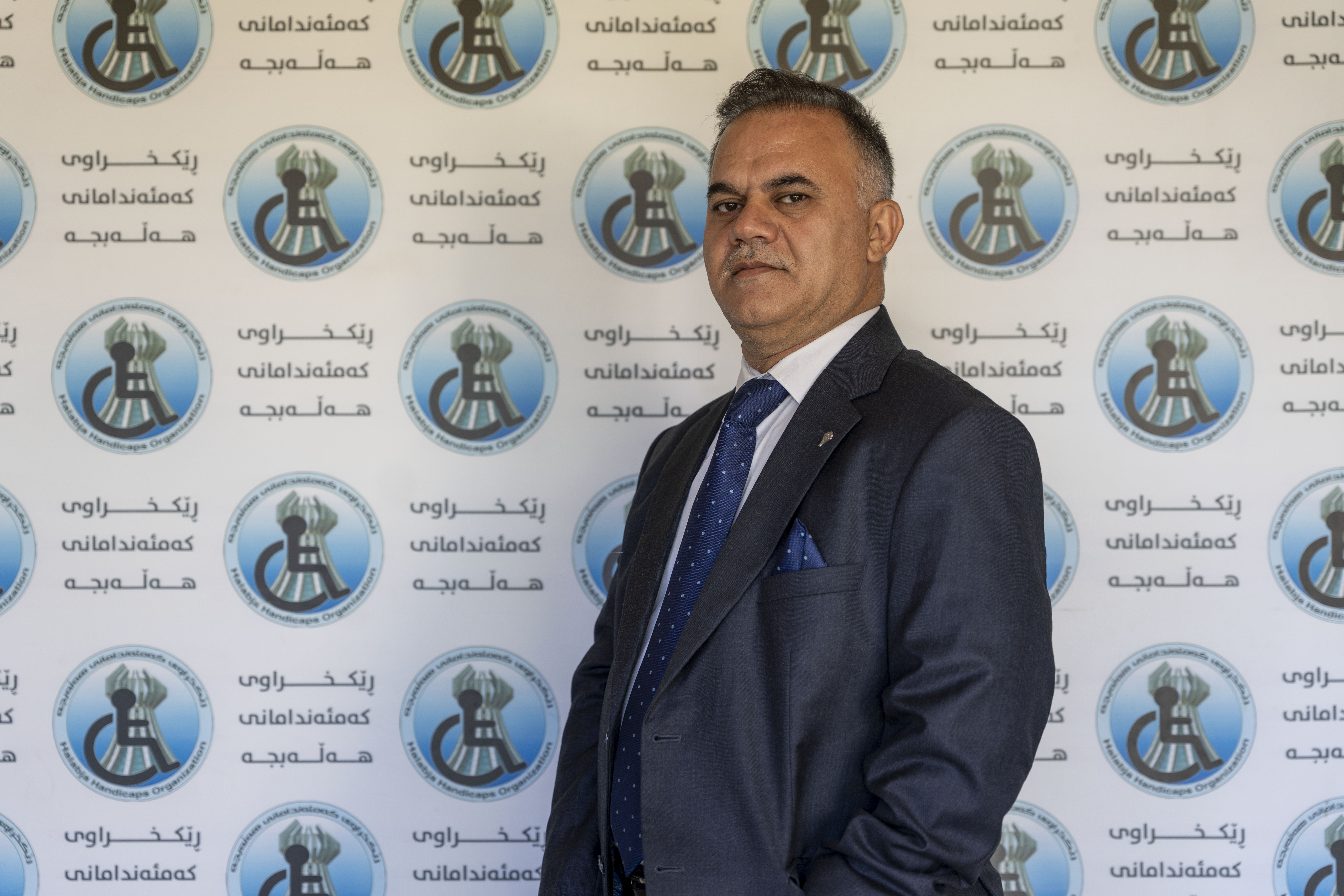 Pukuun pukeutunut mieshenkilö seisoo seinän edessä, joka on peitetty HDO-järjestön logoilla.