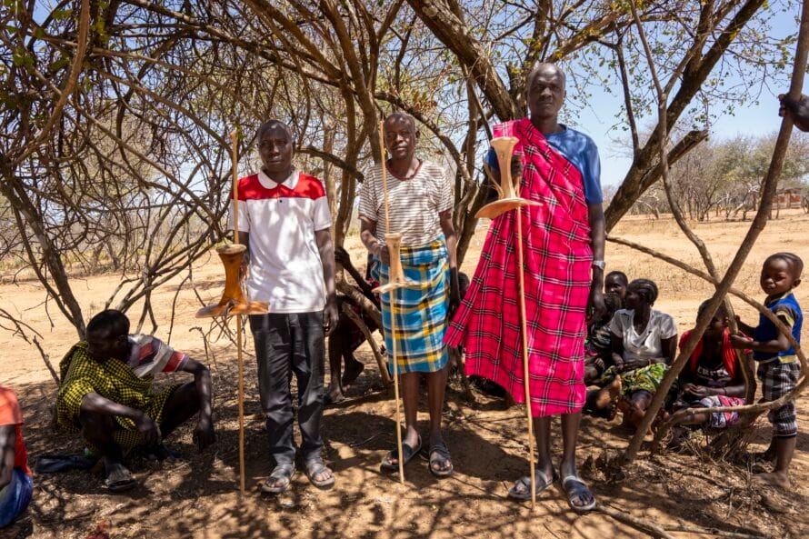 Kolme pokotmiestä seisoo puun alla käsissään perinteiset ng'achara-jakkarat.