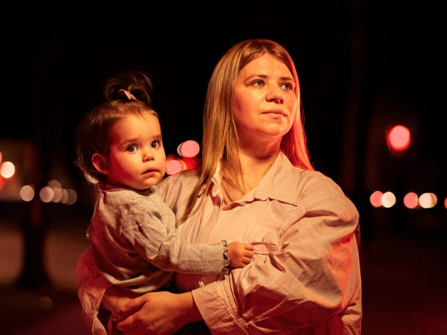 Nuori äiti pieni lapsi sylissään tumman valoja vilkkuvan taustan edessä.