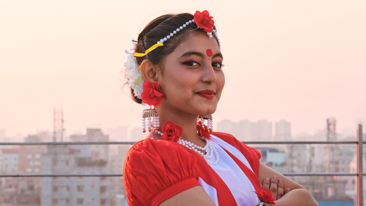 A Bangladeshi girl stands smiling, arms crossed, smiling at the camera, with apartment buildings in the background.