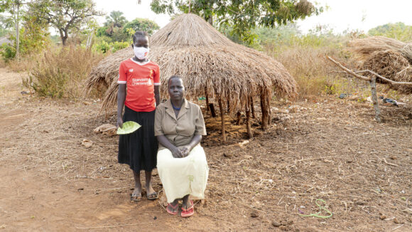 Ugandalainen tyttö seisoo maski päässään ja vieressä istuu nainen katsoen vakavasti eteenpäin, taustalla heinää kuivumassa.