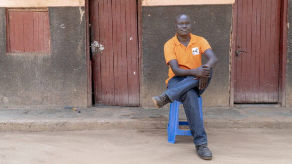 A Ugandan man is sitting outside on a chair, smiling.