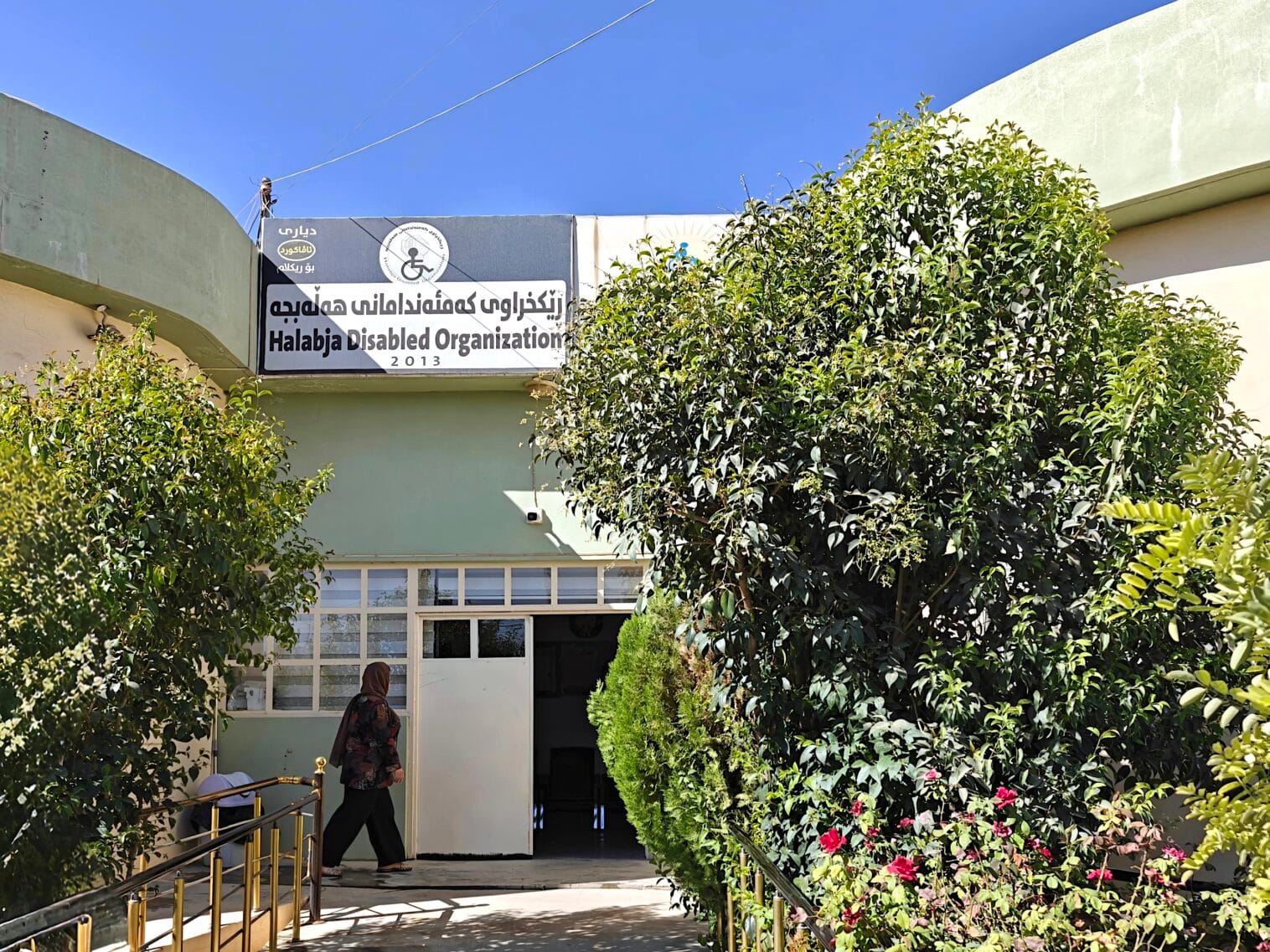 Ulkoapäin kuvattu sisäänkäynti HDO-järjestön toimistorakennukseen. Ovelle johtavan rampin sivuilla kasvaa puskamaisia puita.