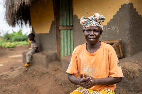 Oranssipaitainen nainen, jolla on päässä huivi, hymyilee kameralle. Taustalla talo.