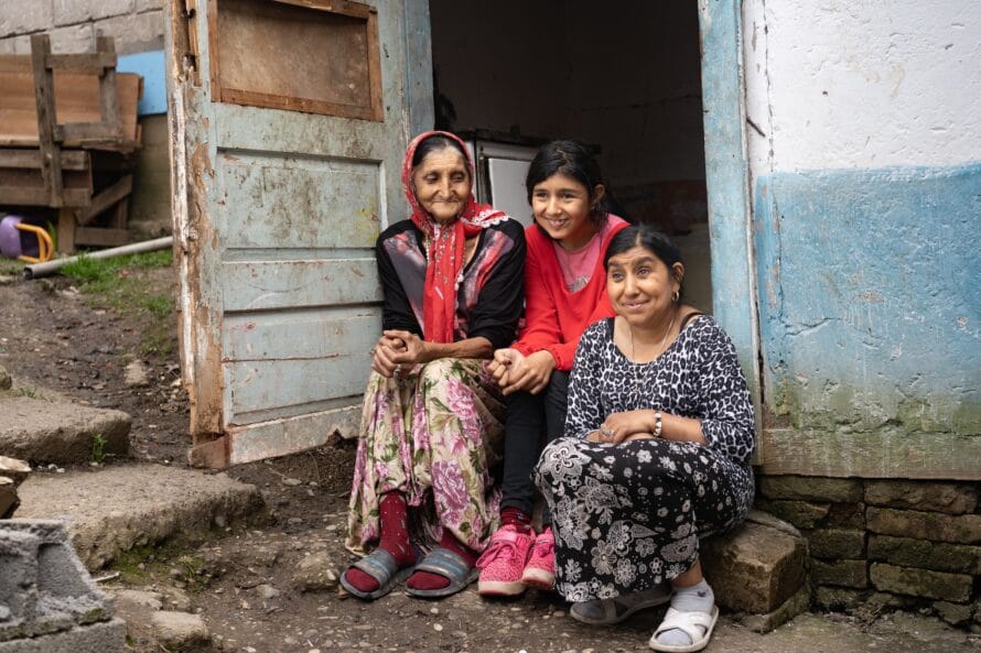 Kolme naista istuu talon portailla, yllään värikkäät vaatteet.