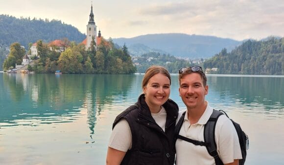 Pariskunta hymyilee kauniin järven rannalla taustallaan kirkkorakennus.