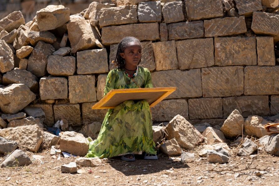 Lapsi istuu romahtaneen koulurakennuksen vieressä, sylissään liitutaulu. 