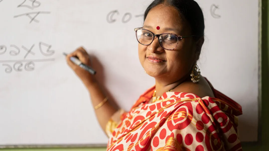 Bangladeshilainen opettajanainen kirjoittaa taululle ja katsoo hymyillen kameraan.