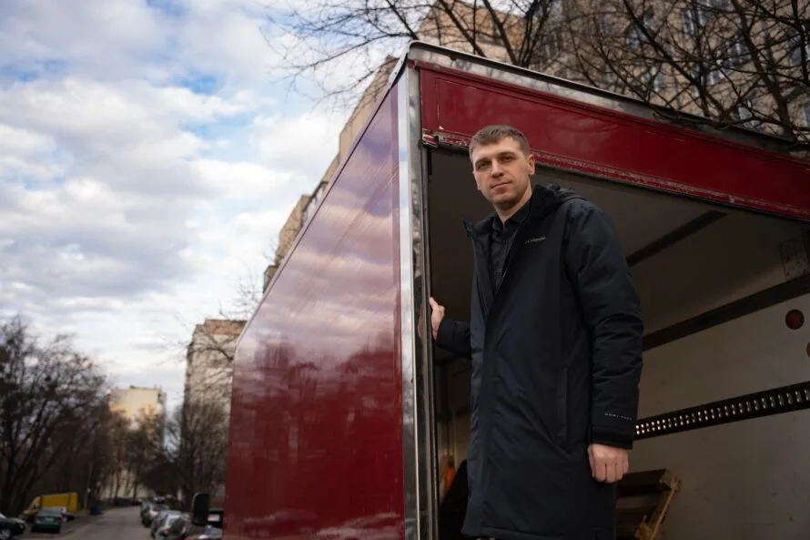 Wearing a dark coat, Marchenko stands at the rear of a red truck, its cargo door open.