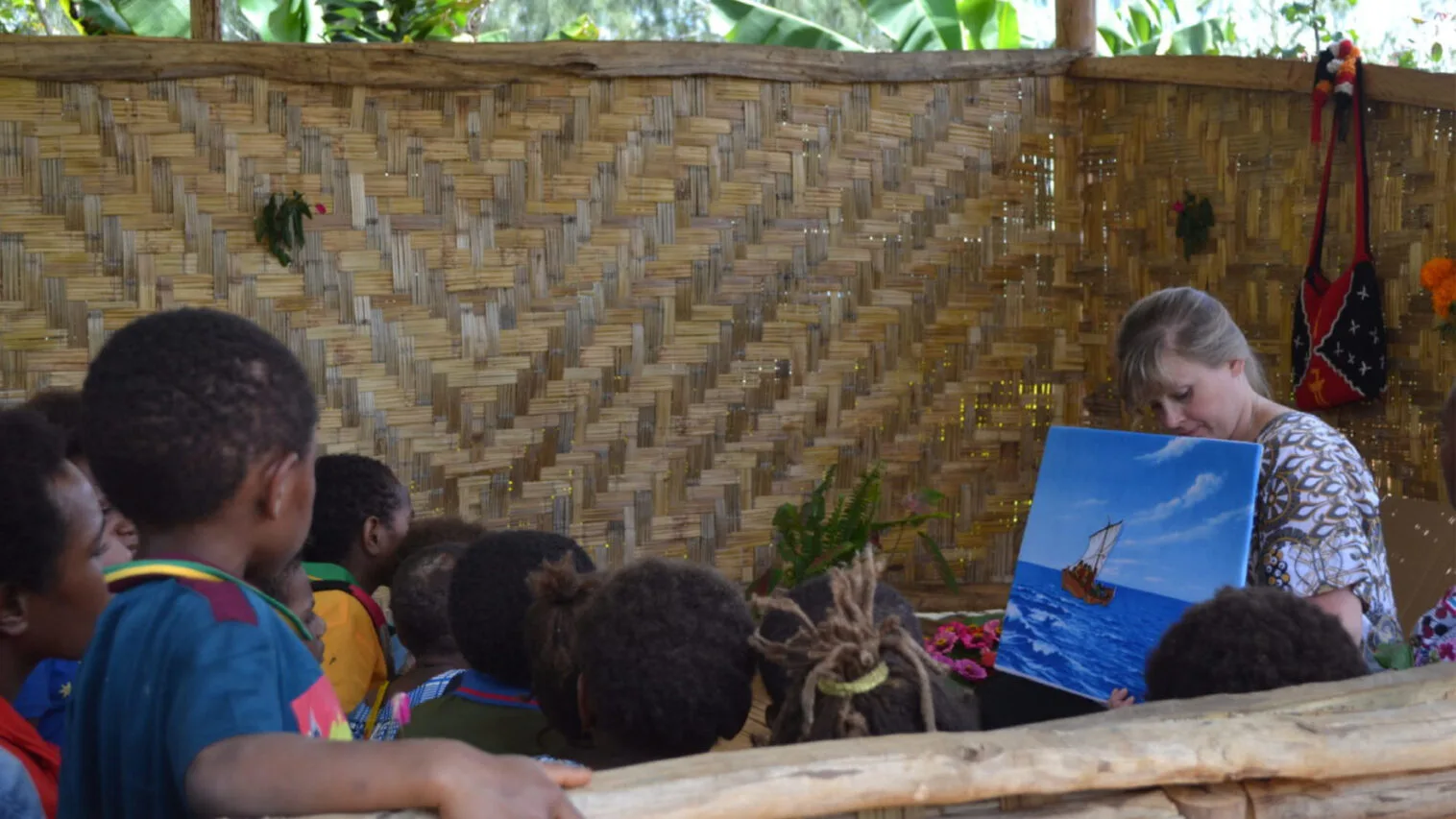 A woman teaching a group of children in Sunday school, holding a painting of a sailboat on a body of water.