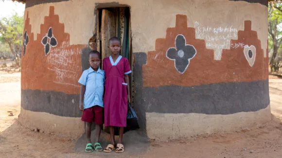 poika- ja tyttölapsi seisovat vierekkäin savimajan edessä ugandalaisessa kylässä.