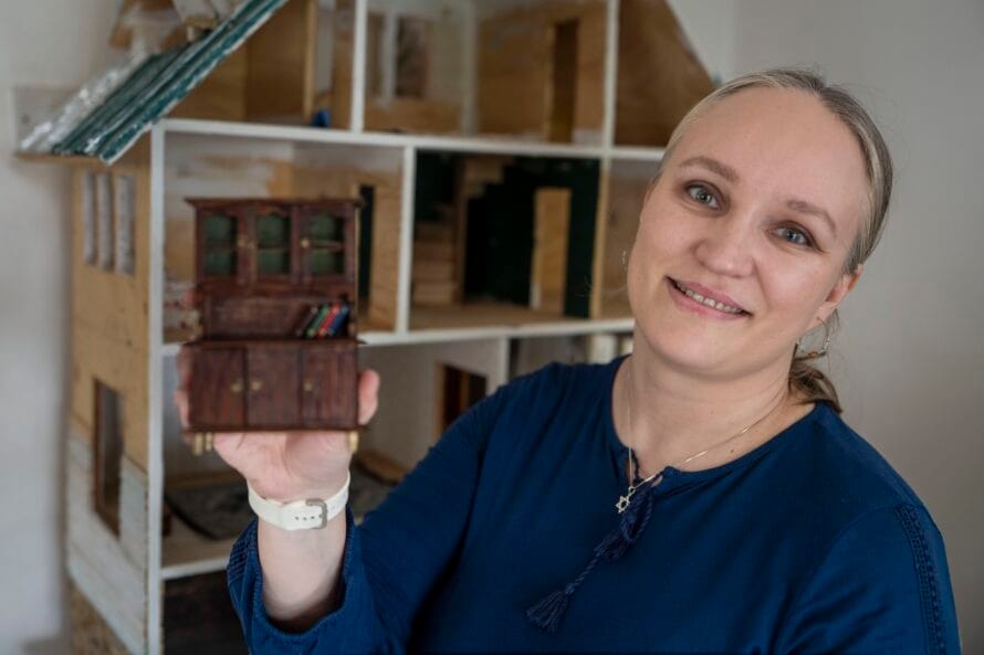 Susanna Sorila (nainen sinisessä paidassa) näyttää kameralle nukkekodin huonekalua, taustalla nukkekoti.