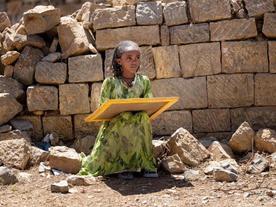 Tyttyö istuu kivellä raunioiden keskellä ja sylissään hänellä on liitutaulu.