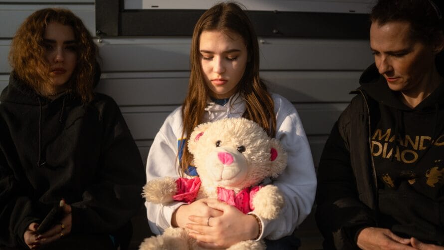 Nuoria naisia istuu ja nojaa rakennusta vasten. Keskimmäisellä nuorella naisella on nalle sylissä.