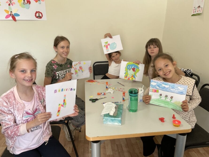 Lapset istuvat pöydän ääressä ja esittelevät värikkäitä piirroksia.