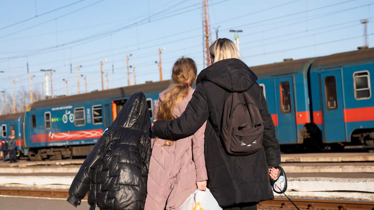 Nainen ja kaksi tyttöä seisovat selin. Taustalla on sininen juna.