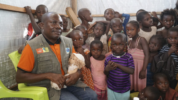 A man is sitting with a doll on his lap and a group of children are standing next to him.