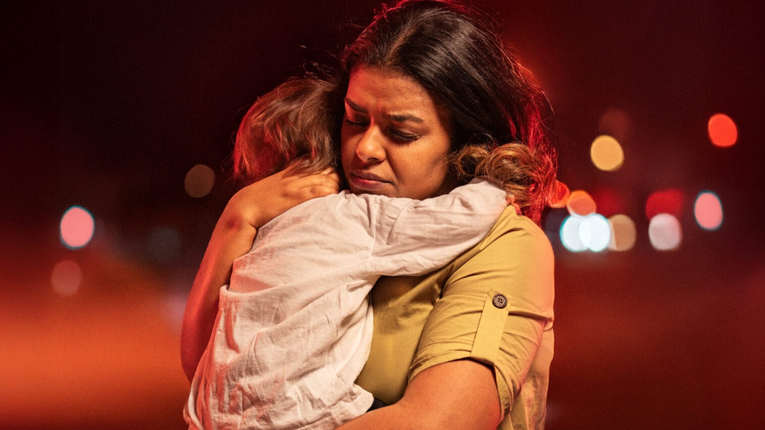 Äiti halaa tiukasti pientä lastaan punertavan valopisteistä koostuvan taustan edessä.