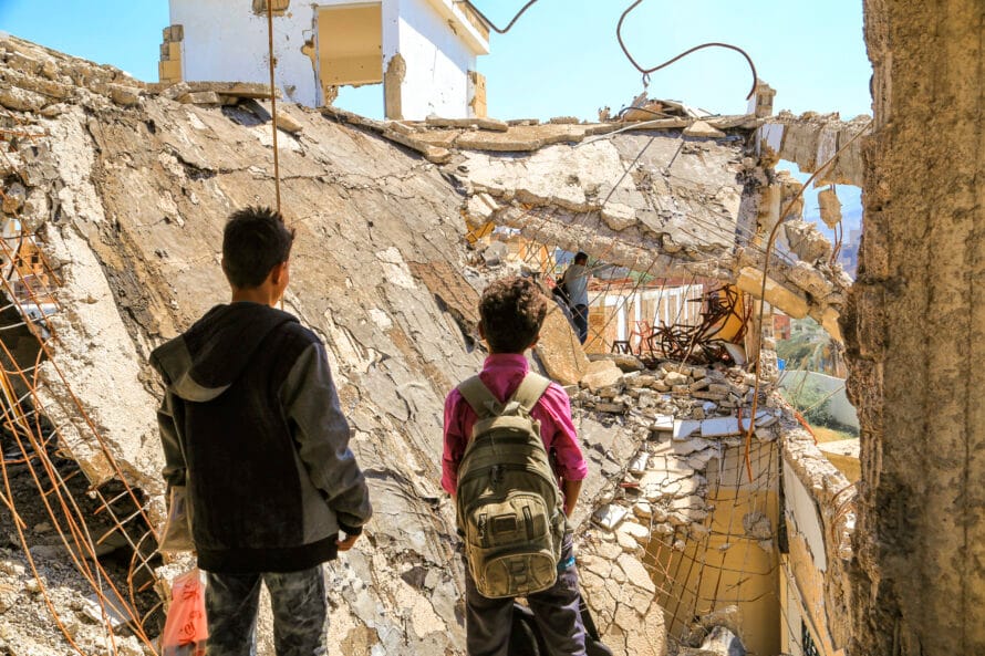 Two boys stand in the middle of the ruins with their backs to the camera.