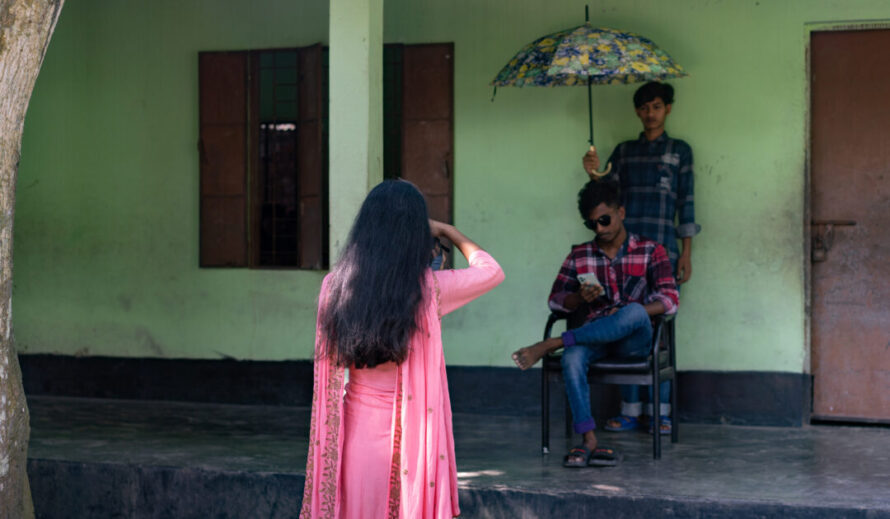 Bangladeshilainen nuori nainen kuvaamassa, taustalla kaksi nuorta miestä, joista toinen istuu puhelin kädessä ja toinen seisoo sateenvarjo kädessä.