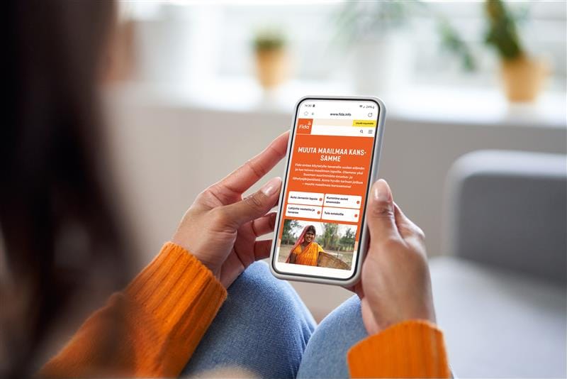 Oranssiin paitaan puetut kädet pitelevät puhelinta, jonka ruudulla näkyy Fidan verkkosivusto.